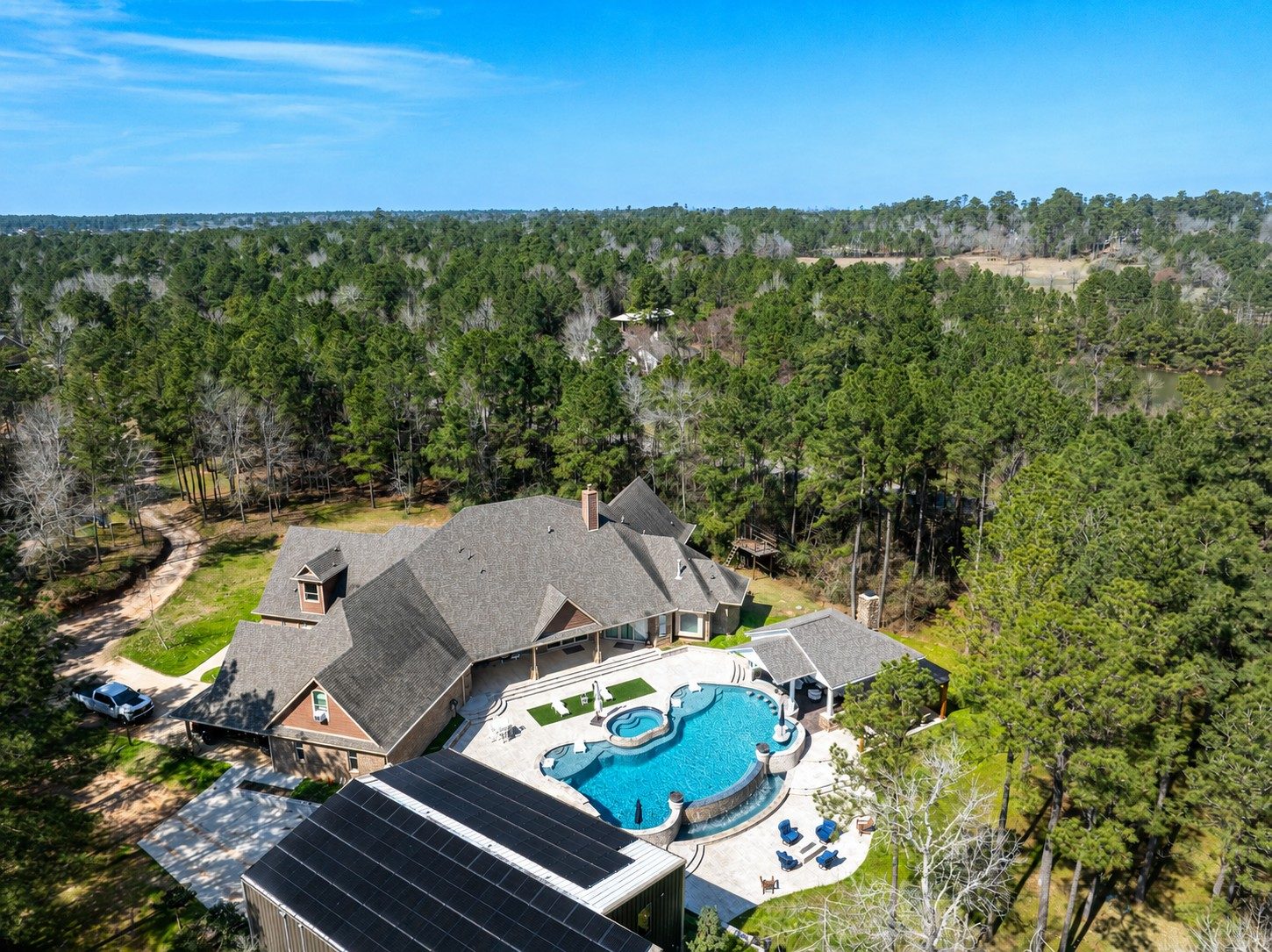 Aerial drone view of a large custom home with a freeform pool, spa, raised sun shelf, and covered patio surrounded by dense pine forest