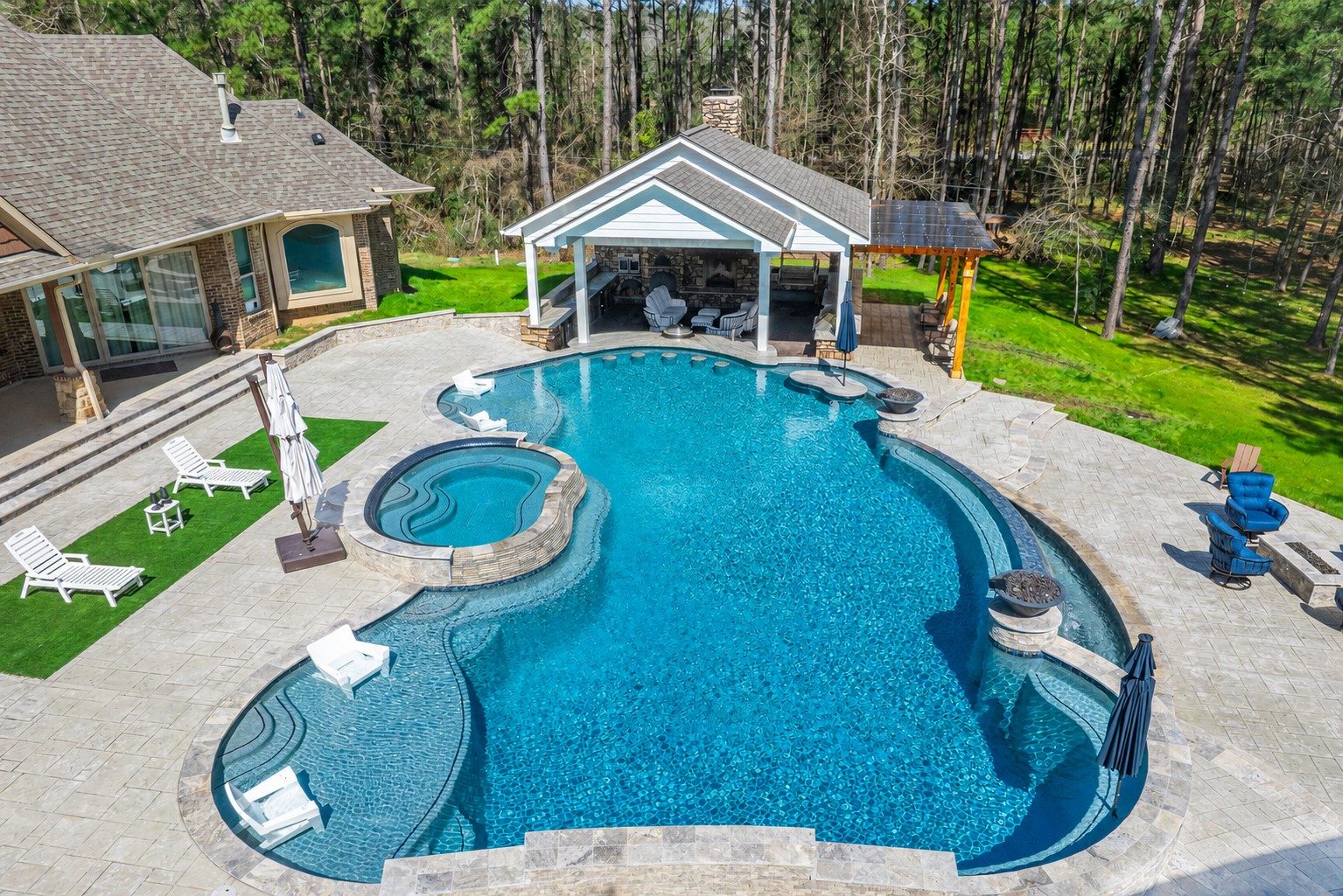 Aerial view of a freeform pool with attached spa, tanning ledge, and paver patio behind a house beside a covered outdoor living pavilion with a fireplace and pergola