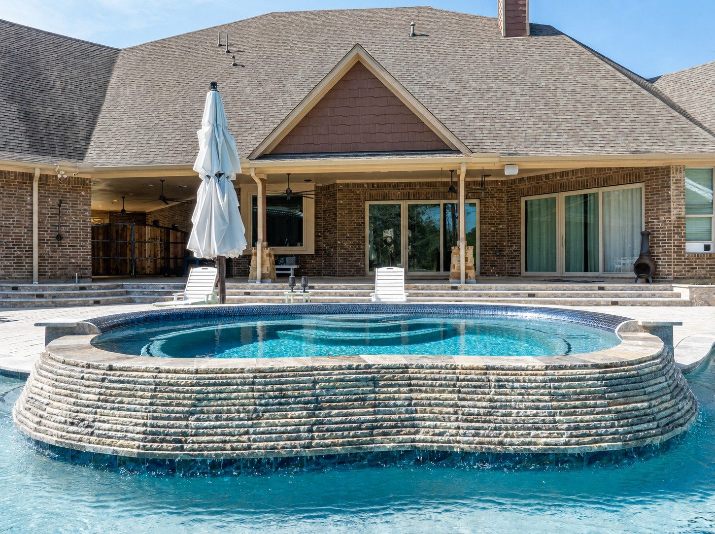 Elevated stone-clad raised spa with sheer-descent waterfalls into a freeform pool in front of a brick house patio with lounge chairs and an umbrella