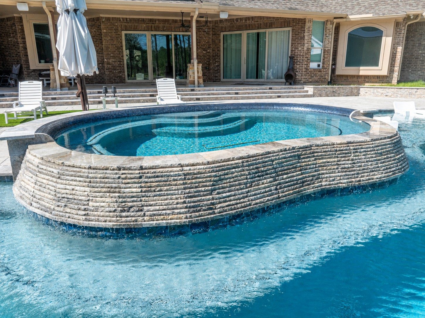 Elevated raised spa with a stacked-stone waterfall wall cascading into a freeform pool behind a brick home patio