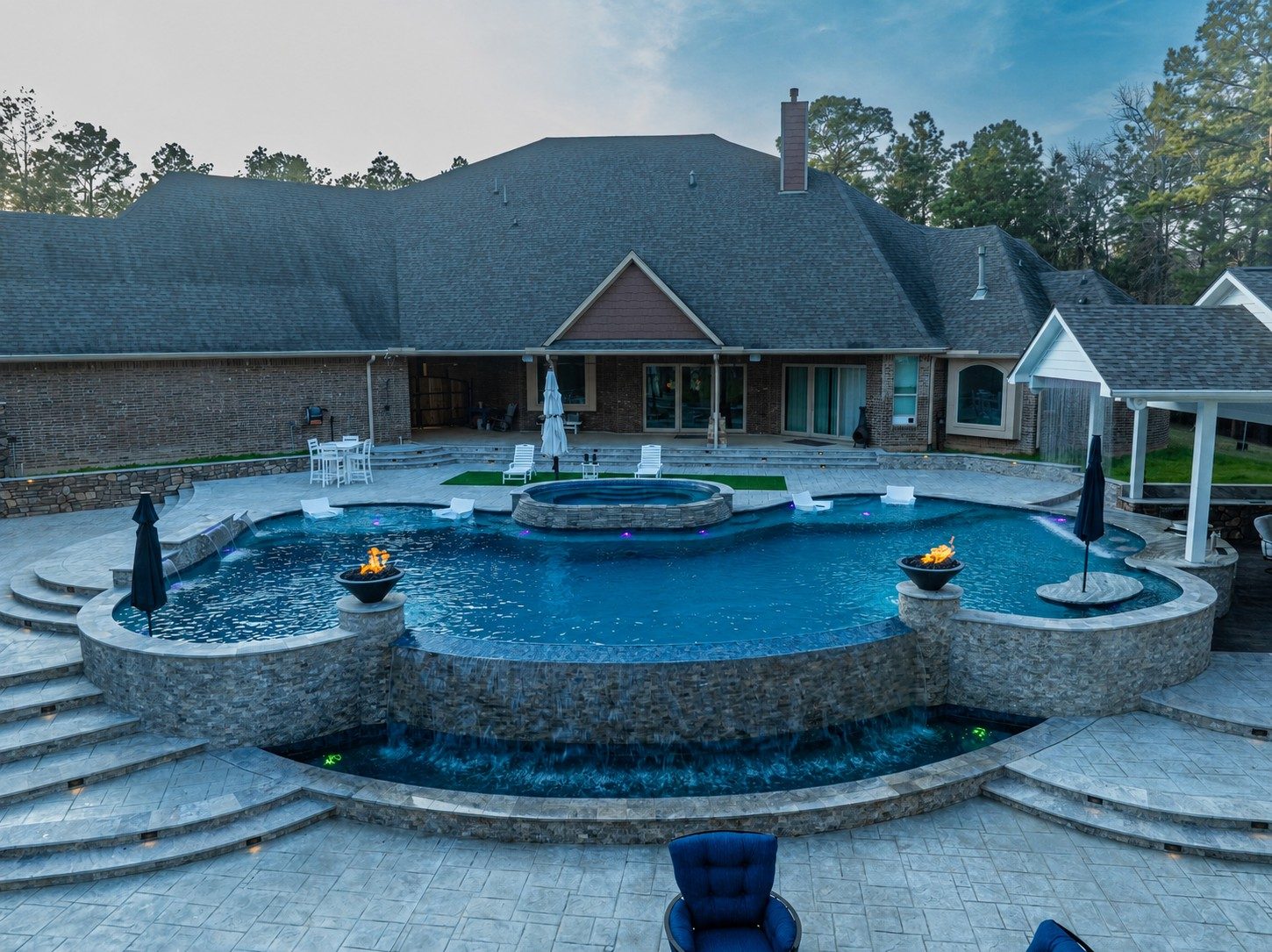Elevated freeform pool with raised spa, twin fire bowls, and vanishing-edge waterfalls spilling into a lower catch pool behind a brick home at dusk
