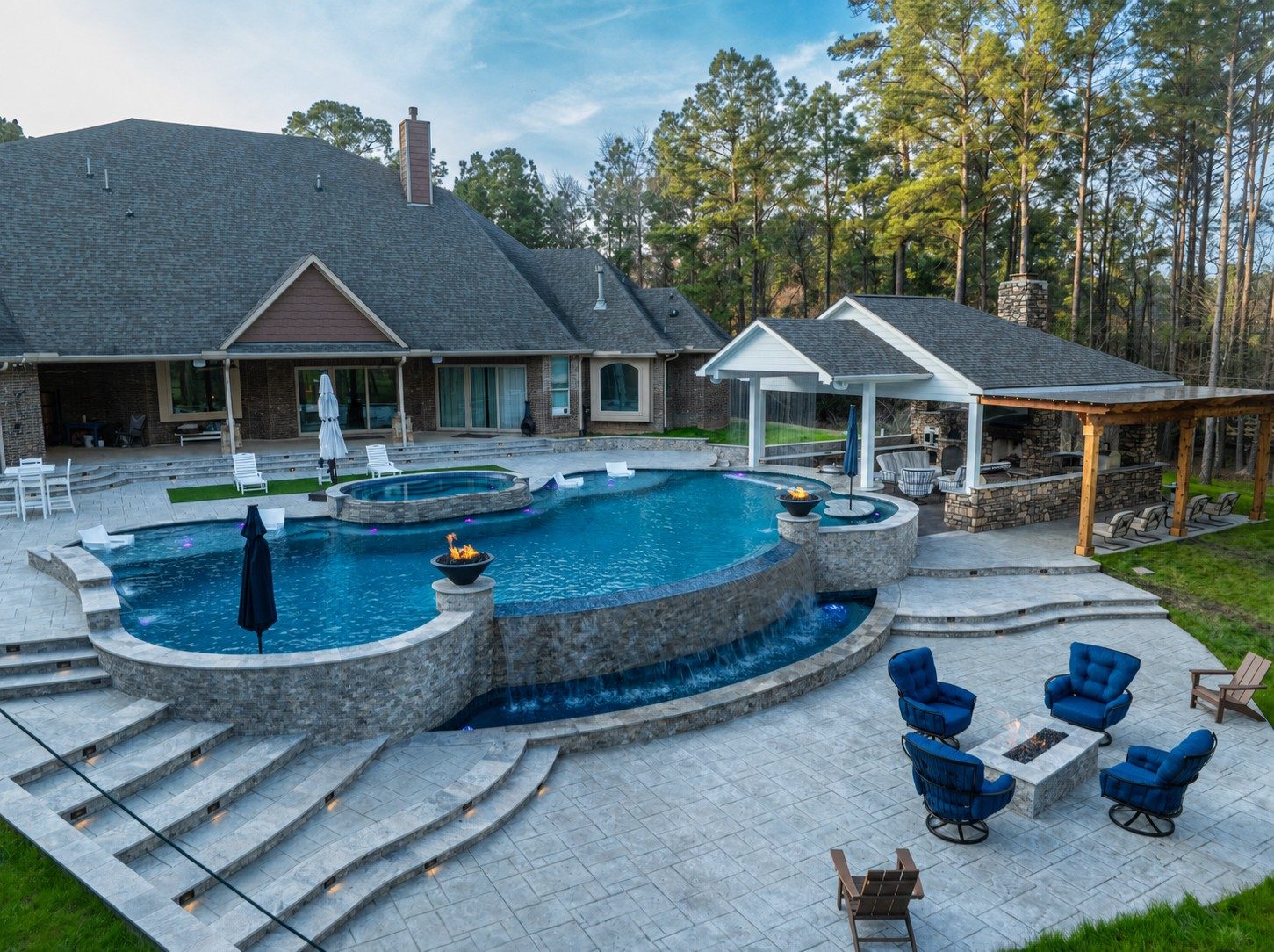 Aerial view of a raised freeform pool and spa with stone waterfall edges, fire bowls, paver patio with blue lounge chairs around a fire pit, and covered outdoor kitchen beside a brick home