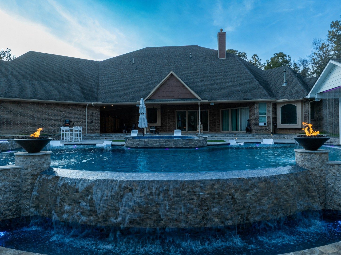 Raised freeform pool with a raised spa cascading water over its tiled edge into a lower catch pool, flanked by two flame bowls in front of a large brick home at dusk