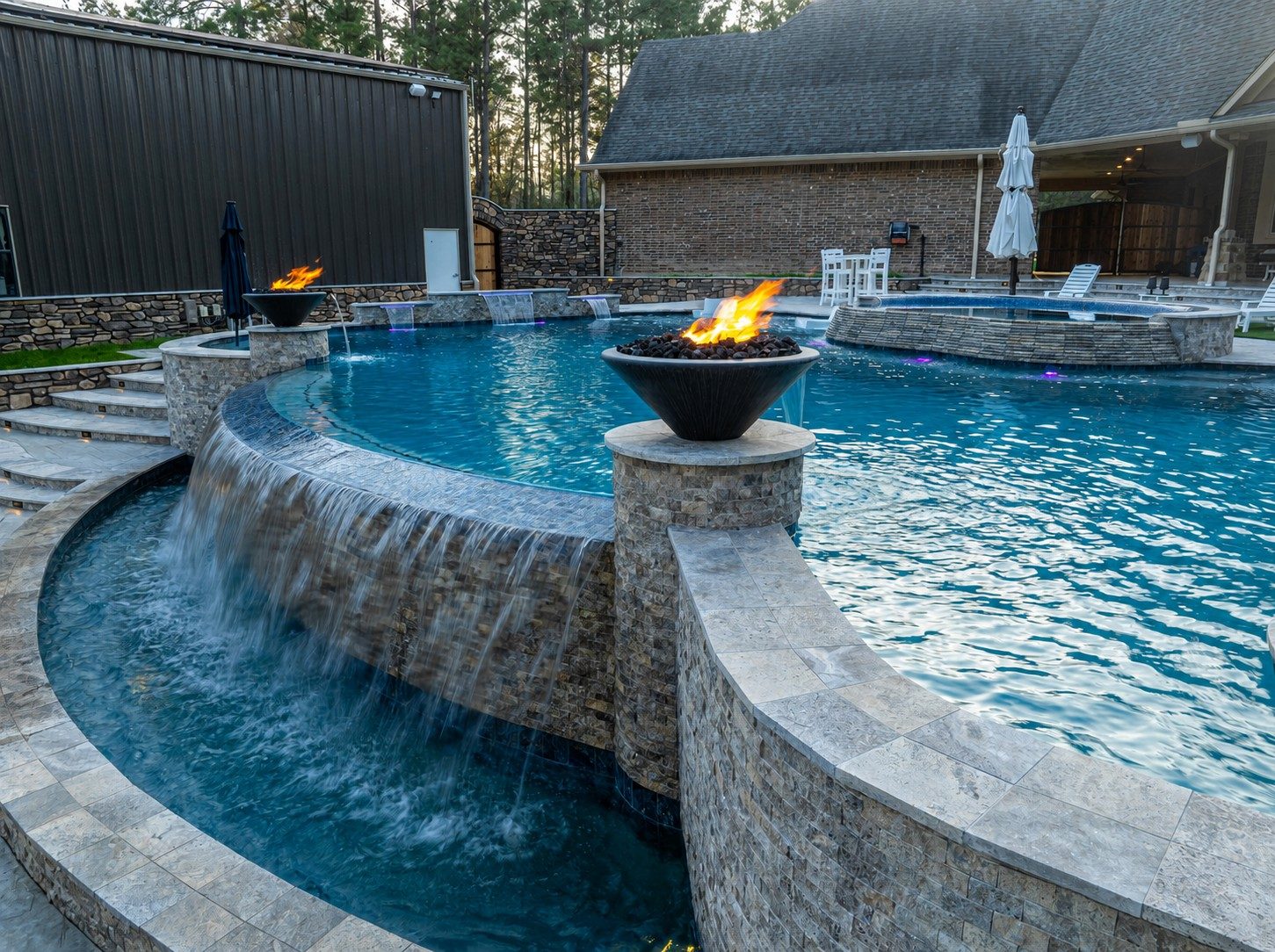 Freeform pool with raised spa cascading over a stone wall past two lit fire bowls into a lower catch basin with a stone-and-metal home in the background at dusk