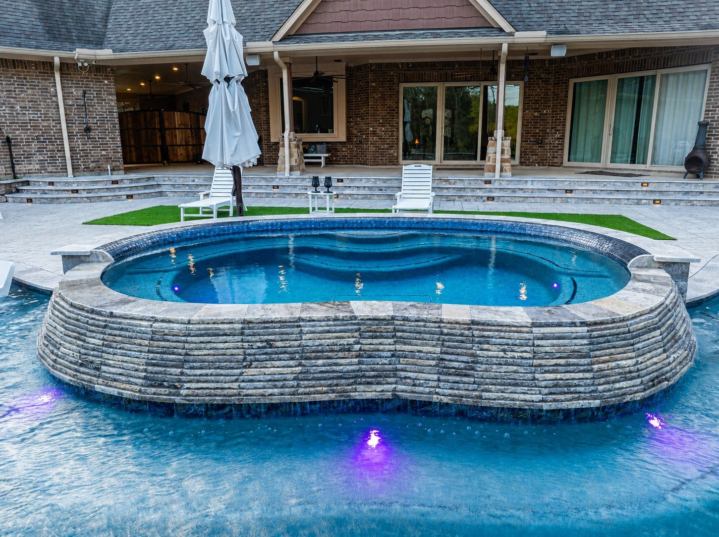 Raised stacked-stone spa with cascading water sheets into a surrounding freeform pool on a travertine patio behind a brick home with purple LED lighting at dusk