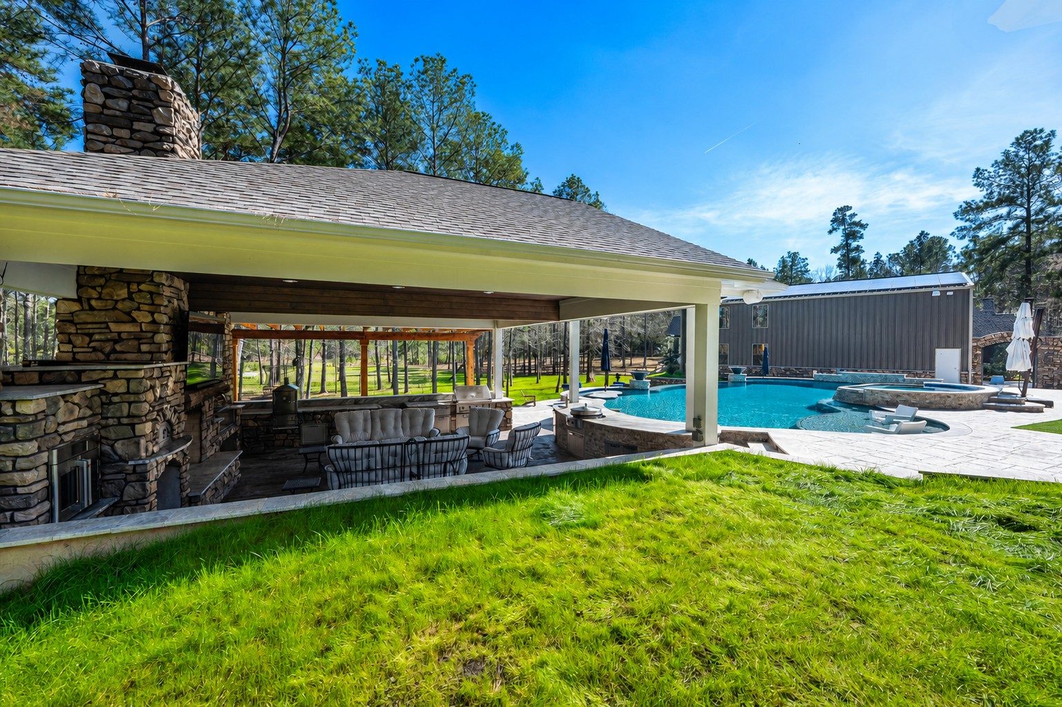 Stone-columned outdoor pavilion with fireplace, kitchen, and lounge seating overlooking a freeform pool and spa surrounded by a paver patio and lawn