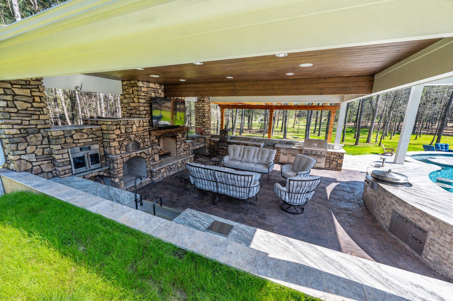Covered outdoor living area with a stone fireplace, mounted TV, wood-fired pizza oven, wicker seating, and a built-in stone kitchen with grill and pool visible alongside