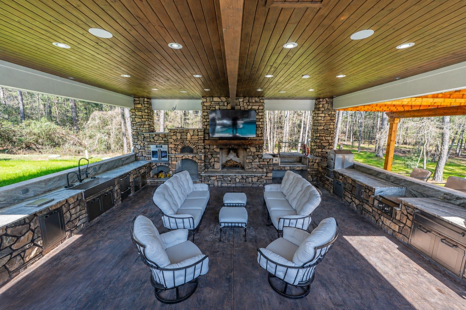 Covered outdoor living pavilion with stone fireplace, mounted TV, wraparound stone kitchen counters, and white cushioned seating on a wood plank floor and ceiling