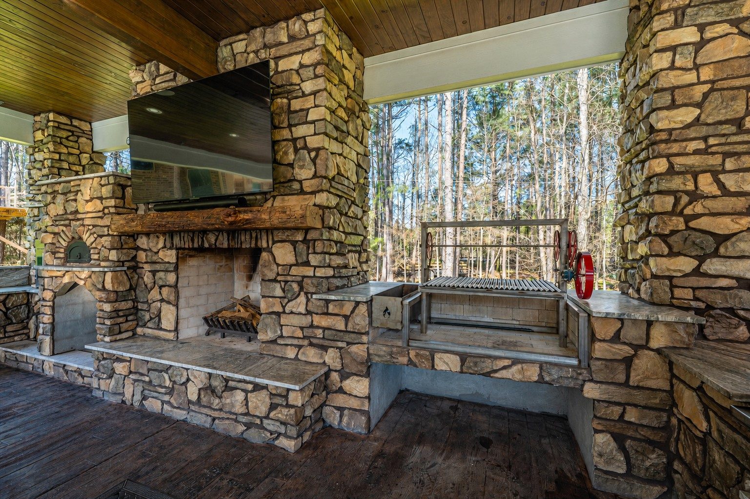 Stone outdoor pavilion with a fireplace, mounted TV, wood-fired pizza oven, and Argentine-style wood grill with wheel-adjustable grates overlooking a wooded backyard