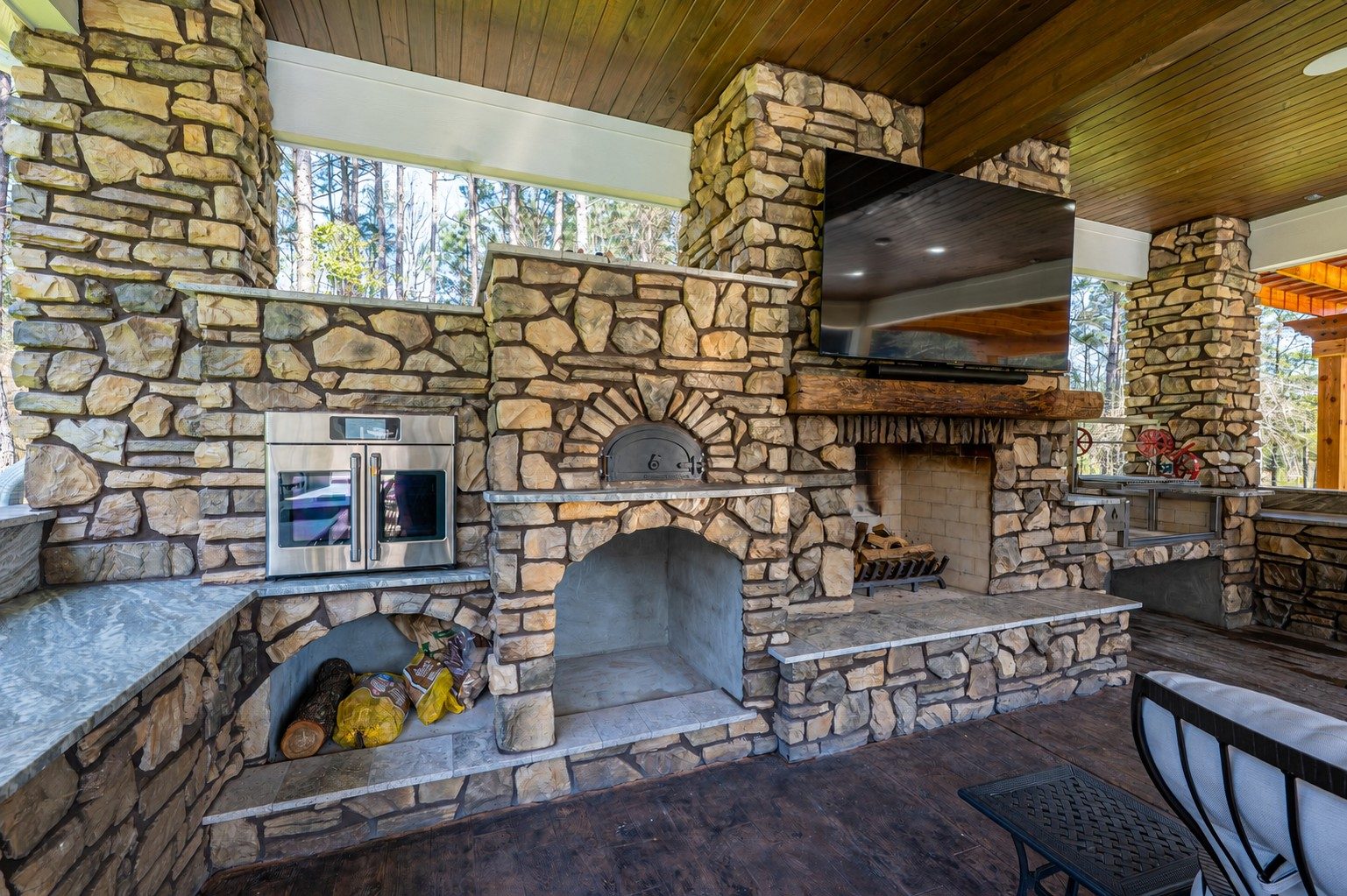 Stone outdoor kitchen and living area with a built-in pizza oven, fireplace with mounted TV, and stainless-steel appliances under a covered wood ceiling