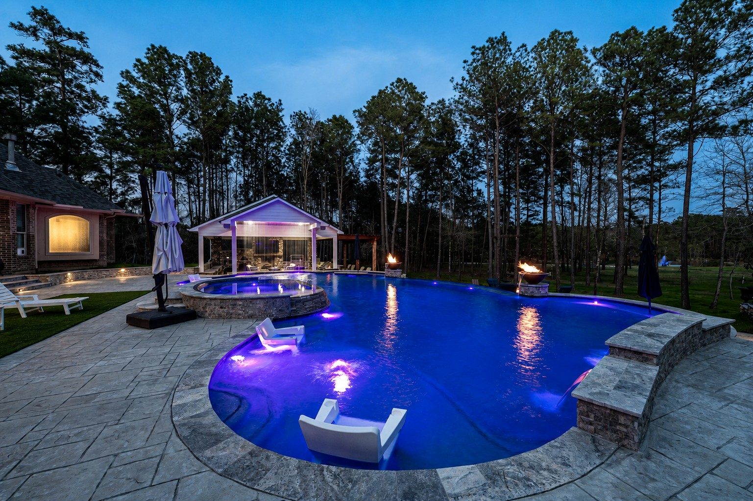 Freeform pool and raised spa glowing with blue LED lighting at dusk beside a pavilion, twin fire bowls, and floating in-pool loungers