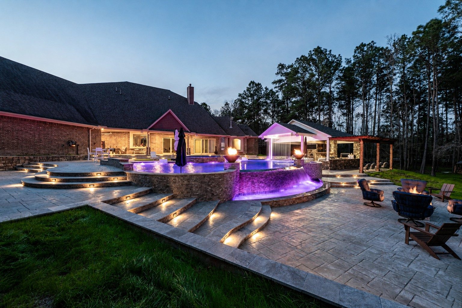 Raised freeform pool and spa with purple LED lighting cascading over a stone waterfall onto a stamped-concrete patio with fire bowls and lounge seating at dusk