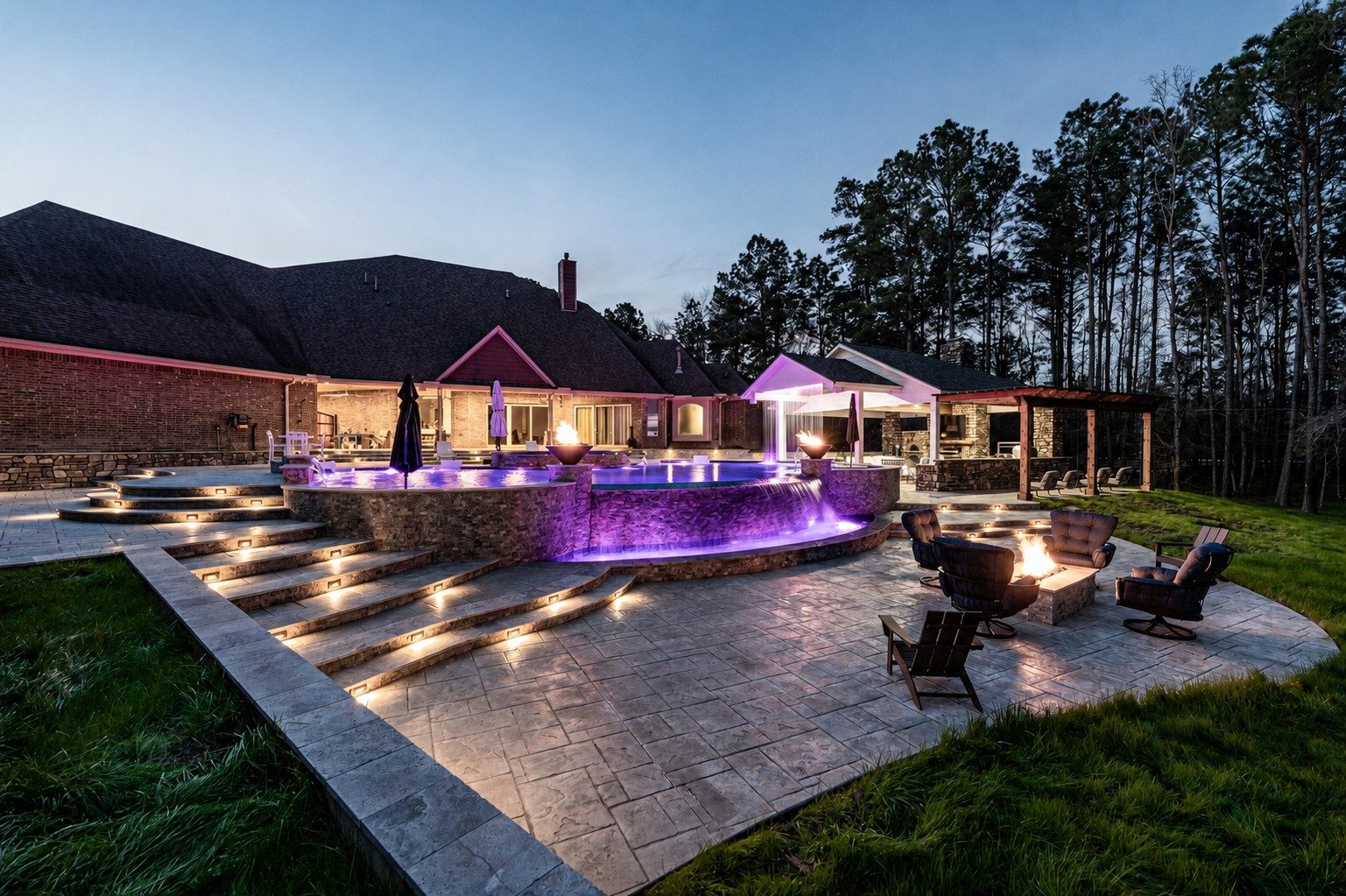 Raised freeform pool and spa glowing with purple LED lighting and a waterfall at dusk beside a stamped-concrete patio with a fire pit and lounge chairs