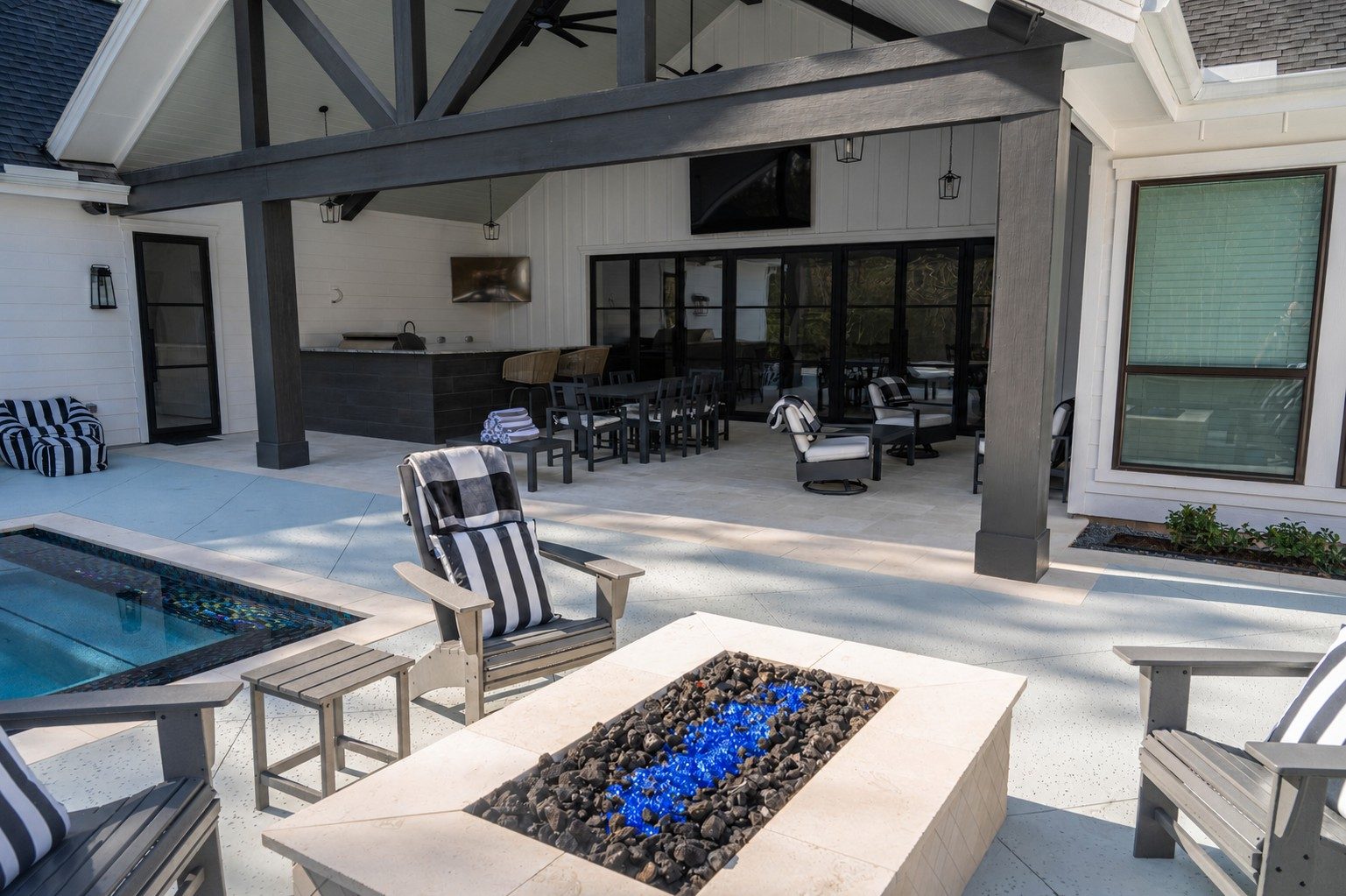 Covered outdoor kitchen and dining pavilion with a deck-level gas fire pit in the foreground, bar stools, dining table, lounge seating, and a TV mounted on the white-board-and-batten wall of the modern farmhouse